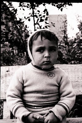 Vintage black-and-white portrait of a young child in a striped sweater with a knitted headscarf, seated outdoors on a wooden ...