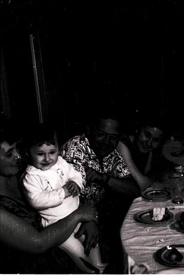 Vintage black-and-white photo of a family gathering at an outdoor table, likely mid-20th century. A child in a white shirt si...