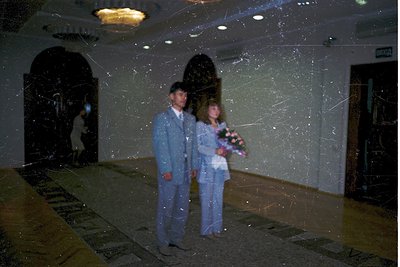 Couple in matching formal wear—light blue suits—posing indoors with a bouquet. Reflective glass walls and ceiling create a mi...
