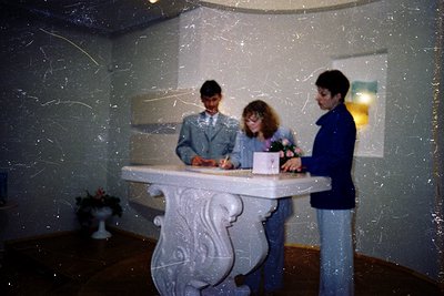 Three individuals signing a ledger at a vintage marble reception desk with ornate carvings, set against textured wallpaper. M...