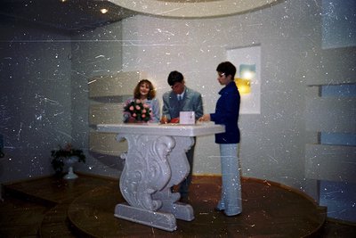 A 1970s-style indoor wedding ceremony with three individuals at a marble altar. The bride in floral dress holds a bouquet, fl...