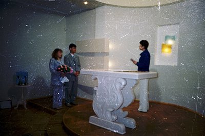 Vintage indoor wedding ceremony with three figures in formal attire. The groom and bride stand beside a white altar adorned w...