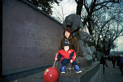 A woman and child pose beside a monumental bronze bear statue on a concrete ledge, holding a red balloon. The bear, seated on...