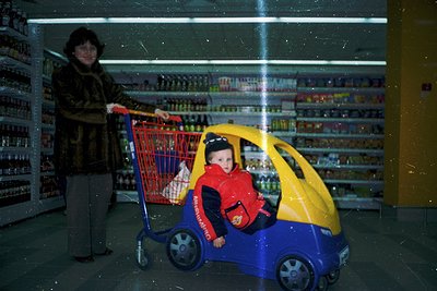A woman in a patterned coat pushes a red shopping cart while a child rides a toy car (blue/yellow) inside. Shelves stocked wi...