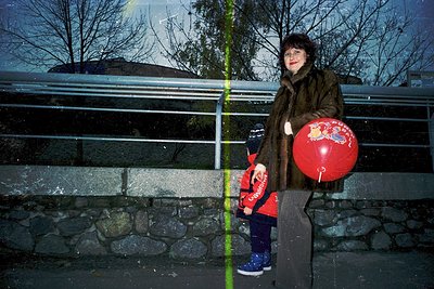 A woman in a brown fur coat poses with a child on a modern bridge railing, holding a red balloon with a white emblem. The sce...