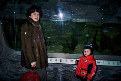 A woman and child stand beside a large indoor aquarium, illuminated by artificial light. The woman wears a dark, textured coa...