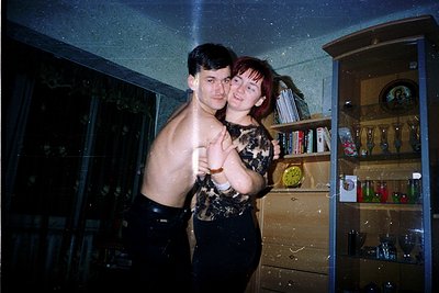 Vintage indoor portrait of two individuals posing indoors, likely late 20th century. The man wears a patterned sleeveless shi...