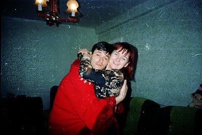 Vintage candid shot of two individuals hugging indoors, likely late 1990s–2000s. Warm lighting from wall sconces illuminates ...