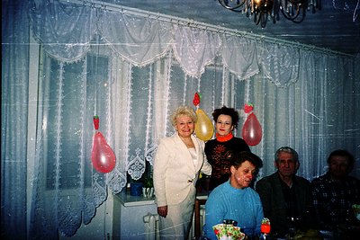 Vintage indoor celebration with three women and a man in a modestly decorated room. The woman in white stands beside a lace c...
