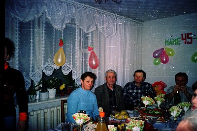 Vintage indoor celebration with four adults seated at a festively decorated table. Decor includes balloon clusters (red, gree...