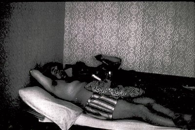 Vintage black-and-white shot of a person reclining on a patterned wallpapered bed, holding a patterned pillow. The geometric ...
