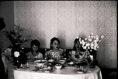 Three women seated at a formal table setting in a mid-century dining room, likely 1960s–1970s. Geometric wallpaper and floral...