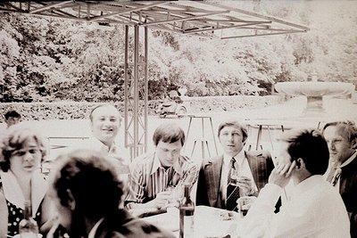 Group of men and women in 1970s-era attire gathered under a metal pavilion, likely at an outdoor event or resort. Formal suit...