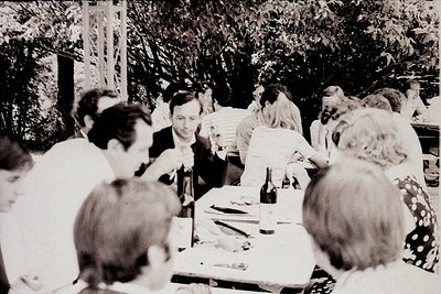 Vintage black-and-white gathering at an outdoor table laden with food, wine, and papers. Mid-20th century attire suggests 195...