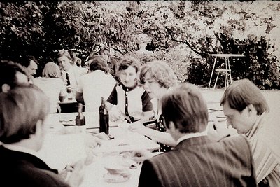 Mid-20th century outdoor gathering at a long dining table under leafy trees. Formal attire suggests a professional or celebra...