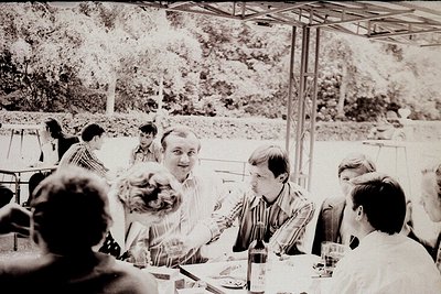Group of 10+ casually dressed individuals seated at a covered outdoor dining area, likely mid-20th century (1960s–1970s). Woo...