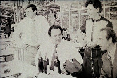 Four men in formal attire—one in a white lab coat, another in a suit with a striped tie, and two in dark suits—gather around ...