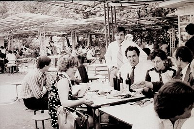 Black-and-white snapshot of an outdoor café or restaurant in the 1970s, featuring a lively group dining under white canopies....