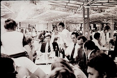 Indoor social gathering in a covered dining area, likely a 1970s European café or restaurant. Men in suits and women in dress...