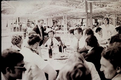 Mid-century indoor dining hall with long tables, glass partitions, and fluorescent lighting. Groups of men in suits and white...
