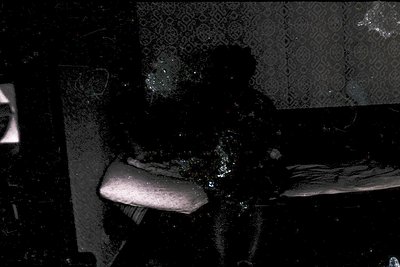 Vintage black-and-white photo of a seated individual in a dimly lit interior, likely a 1950s–1960s living room. Patterned wal...