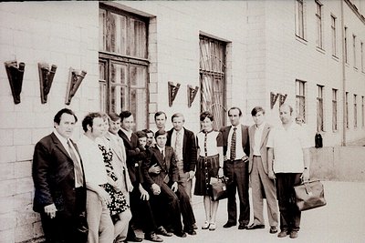 Group portrait in front of a Soviet-era industrial building, likely 1960s–1980s. Ten individuals in formal attire—men in suit...