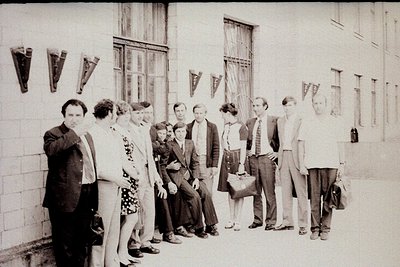 Black-and-white group photo outside a Soviet-era institutional building, featuring 13 individuals in 1960s–70s formal attire....