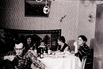Family gathering at a festive table, likely Christmas ( ). Indoor setting with patterned wallpaper and a framed clock. Adults...