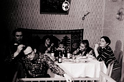 Family gathering at a table set with vintage tableware, likely mid-20th century. Six individuals seated around a round table ...