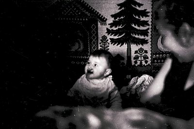 Vintage black-and-white portrait of a young child seated indoors, surrounded by intricate embroidered wall hangings depicting...