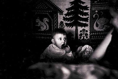 Black-and-white photograph of a young child seated indoors, holding a stuffed animal. Intricate embroidered wall hangings fea...