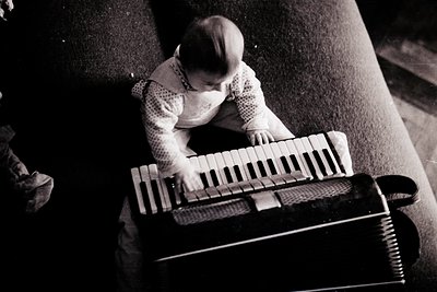 Young child playing a vintage accordion in an indoor setting, mid-20th century. The child’s knit sweater and focused expressi...
