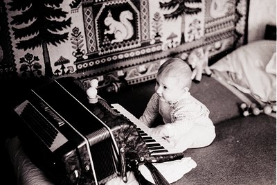 Vintage black-and-white photo of an infant playing a vintage accordion on a couch, surrounded by ornate wallpaper with floral...