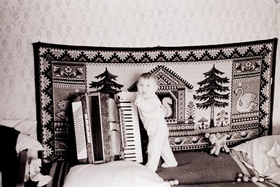 Vintage black-and-white photo of a toddler standing on a ladder beside a decorative quilt with pine trees, swans, and geometr...