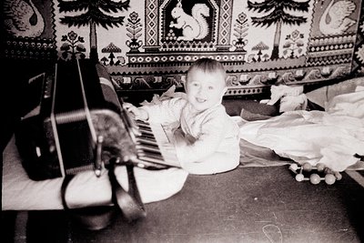 Mid-20th-century black-and-white photo of a toddler playing a grand piano in a cozy, patterned room. Ornate wallpaper feature...