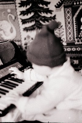 Vintage black-and-white photo of a child playing a vintage upright piano, hands positioned on keys. Patterned wallpaper with ...