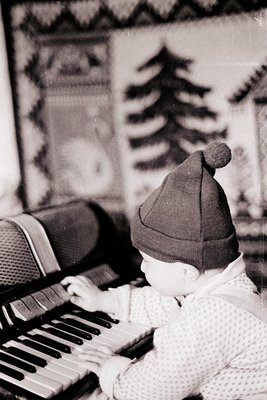 Vintage black-and-white photo of a toddler wearing a knit Santa hat playing a vintage upright piano. Patterned wallpaper with...