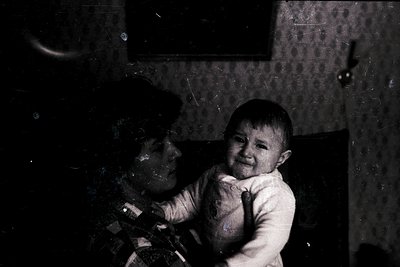 Vintage black-and-white photo of an infant held by an adult in a dimly lit indoor setting, likely mid-20th century. The child...