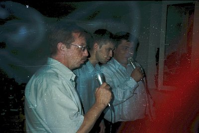 Vintage photo of three men speaking into microphones, likely at a press conference or event. Blurred reflections and low ligh...