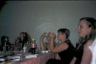 Vintage indoor party scene with blurred motion effects, likely from a 1980s–1990s film. Three adults at a table—one holding a...