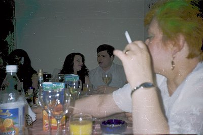 Vintage indoor gathering with blurred faces, featuring a woman in a light-colored blouse smoking. Table laden with empty bott...