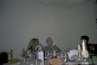 Vintage indoor celebration scene with three blurred figures seated at a table. Decor includes a decorative figurine, champagn...