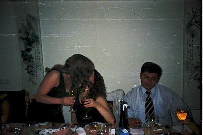 Vintage indoor gathering featuring three adults at a table with dim lighting. Woman in black dress leans over to kiss another...
