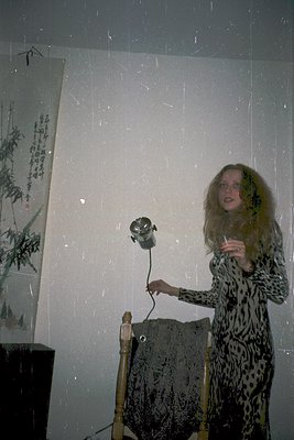 Vintage portrait of a woman in a leopard-print dress holding a cigarette, standing beside a vintage camera tripod and a drape...