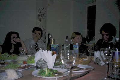 Vintage indoor gathering at a rectangular table with white tablecloth, featuring four adults seated. Centerpiece includes dec...