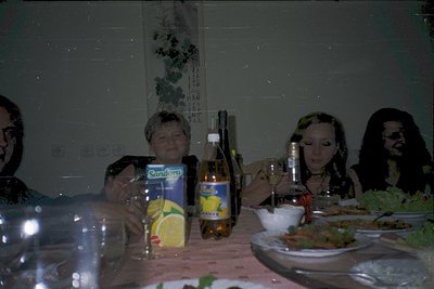 Vintage indoor gathering featuring four individuals seated at a wooden table. Centerpiece includes a bottle of beer, a carton...
