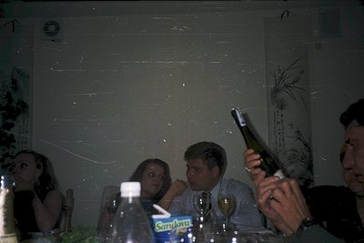 Vintage indoor gathering with blurred lighting, featuring three adults seated at a table. One man raises a bottle, likely cha...