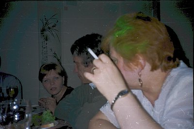 Vintage indoor portrait showing three adults seated at a table, likely mid-1990s. Blurred foreground subject smokes, while ot...