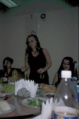 Vintage-style indoor banquet with three women in 1970s-inspired glamour attire. Centered: woman in black sleeveless dress wit...