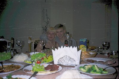 Vintage indoor celebration with two smiling individuals seated at a long table adorned with festive tableware. Plates display...